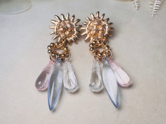 A pair of dangling statement earrings with textured gold sunburst tops and three teardrop-shaped clear and pale pink glass drops, displayed on a light beige surface.