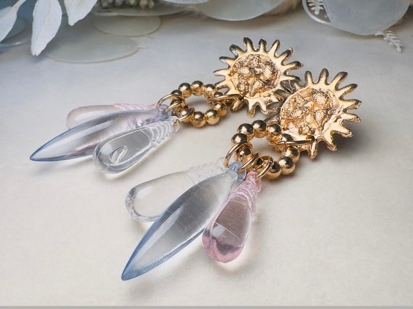 A pair of gold-toned dangle earrings with sunburst-shaped studs, small gold beads, and clear teardrop-shaped crystal drops, photographed on a light neutral surface with soft decorative elements in the background.