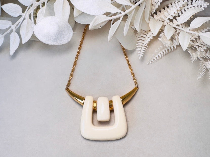 A cream-colored geometric pendant necklace with a gold chain, displayed on a light gray background with white decorative foliage.