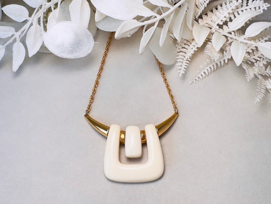 A cream-colored geometric pendant necklace with a gold chain, displayed on a light gray background with white decorative foliage.