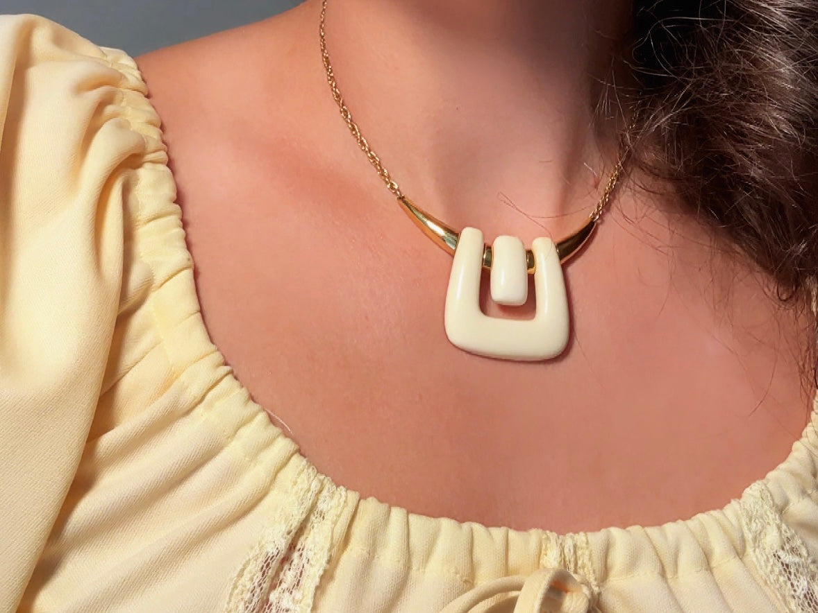 A close-up of a person wearing a cream-colored geometric pendant necklace with a gold chain and a pale yellow blouse.