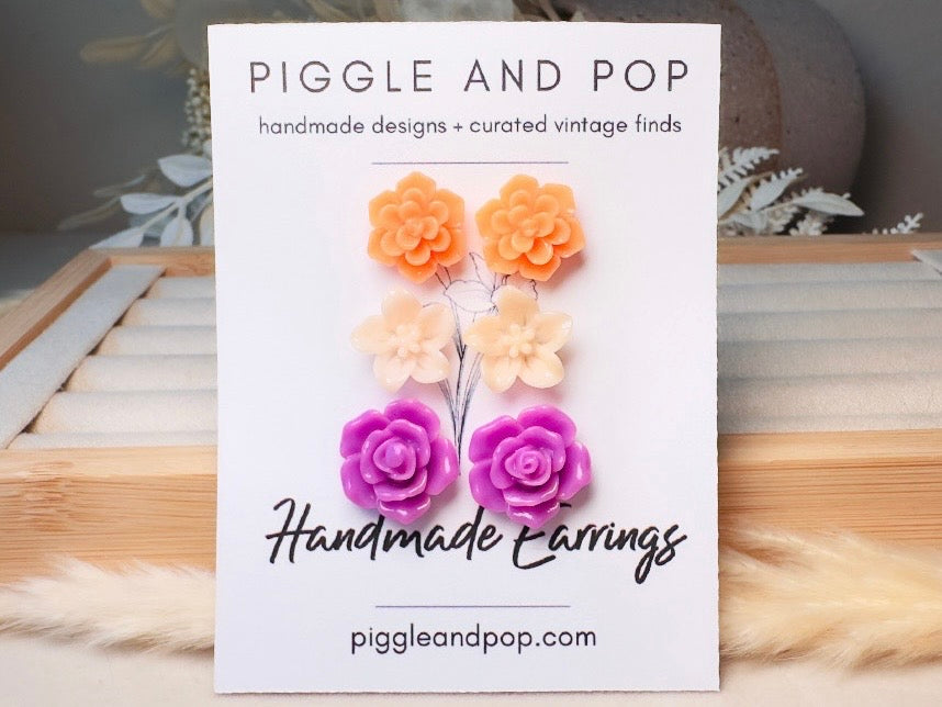 A white display card for Piggle and Pop handmade earrings, featuring four flower-shaped earrings in orange, peach, and purple. The card is displayed on a wooden surface with a soft beige faux fur accent.