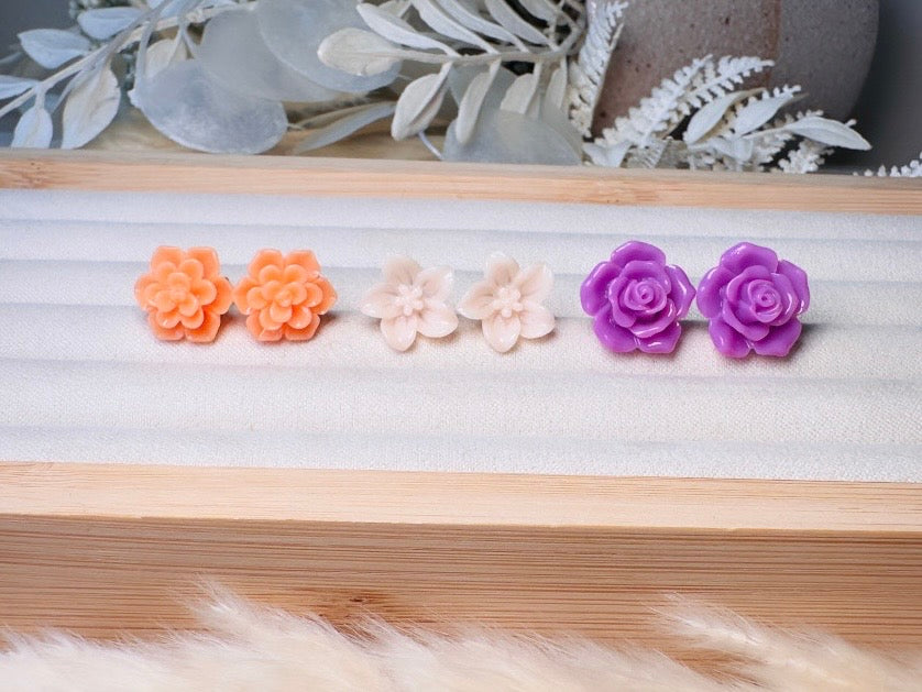 Four pairs of small flower-shaped stud earrings arranged on a light wood tray. The pairs are orange, pale pink, and purple, with decorative dried leaves in the background.