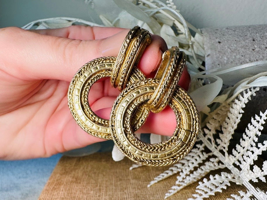 These vintage gold door knocker earrings are absolutely fabulous! Gold tone door knocker style hoops &nbsp;are chock full of interesting details. These earrings feature 3 rows of textured raised concentric hoops- the outer rings are a gold roped pattern and the center is rows of rectangle panels. These earrings are such a classic Etruscan pair- bold and dramatic! In great vintage condition -no major flaws to note- please see pics. Clip on style!

Dimensions: 1.5" x 1.25"&nbsp;