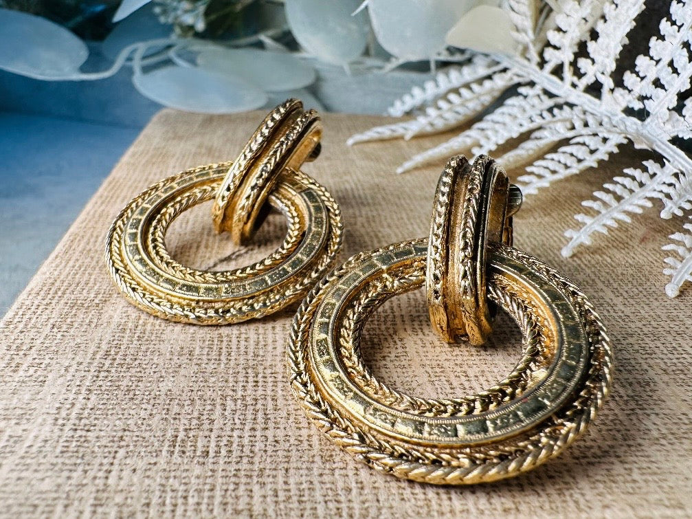 These vintage gold door knocker earrings are absolutely fabulous! Gold tone door knocker style hoops &nbsp;are chock full of interesting details. These earrings feature 3 rows of textured raised concentric hoops- the outer rings are a gold roped pattern and the center is rows of rectangle panels. These earrings are such a classic Etruscan pair- bold and dramatic! In great vintage condition -no major flaws to note- please see pics. Clip on style!

Dimensions: 1.5" x 1.25"&nbsp;