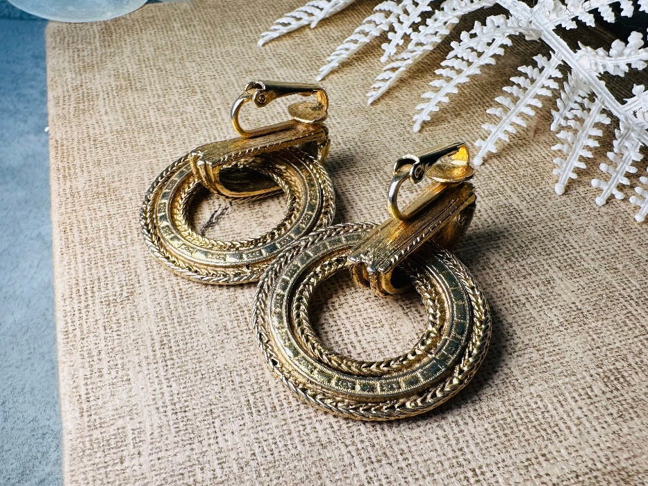 These vintage gold door knocker earrings are absolutely fabulous! Gold tone door knocker style hoops &nbsp;are chock full of interesting details. These earrings feature 3 rows of textured raised concentric hoops- the outer rings are a gold roped pattern and the center is rows of rectangle panels. These earrings are such a classic Etruscan pair- bold and dramatic! In great vintage condition -no major flaws to note- please see pics. Clip on style!

Dimensions: 1.5" x 1.25"&nbsp;