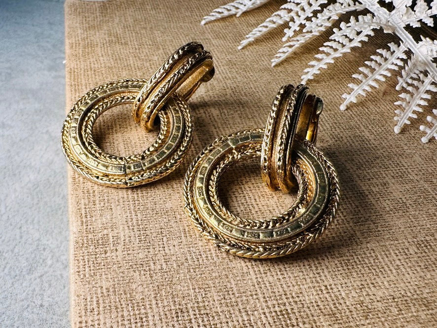These vintage gold door knocker earrings are absolutely fabulous! Gold tone door knocker style hoops &nbsp;are chock full of interesting details. These earrings feature 3 rows of textured raised concentric hoops- the outer rings are a gold roped pattern and the center is rows of rectangle panels. These earrings are such a classic Etruscan pair- bold and dramatic! In great vintage condition -no major flaws to note- please see pics. Clip on style!

Dimensions: 1.5" x 1.25"&nbsp;