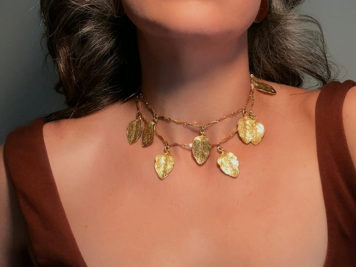 A close-up of a model wearing a gold leaf necklace. The necklace features multiple textured, leaf-shaped pendants on a delicate chain, styled with a brown top against a neutral gray background.