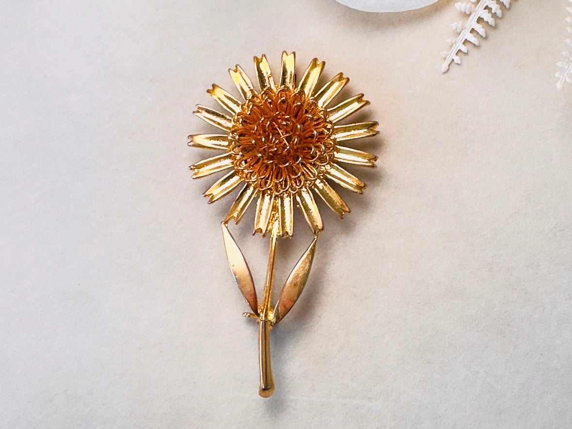 A gold-toned metal brooch shaped like a sunflower, featuring a textured center and radiating petals, displayed on a light beige textured surface.