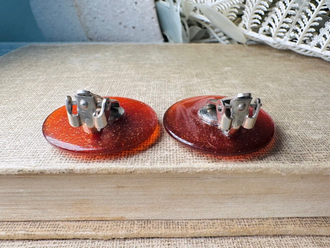 These vintage lucite button earrings from Piggle and Pop are such a gorgeous color- vibrant copper with flecks of fine gold glitter. These retro clip ons are from the 1960s. A fun party pair, these confetti earrings were wildly popular at the time and a fun statement pair to rock in modern day. Lightweight for their size, a round slightly domed coin shape and easy to wear in clip on style. They catch the light beautifully and can appear a deep reddish copper shade to a glowing sunset hue. 
