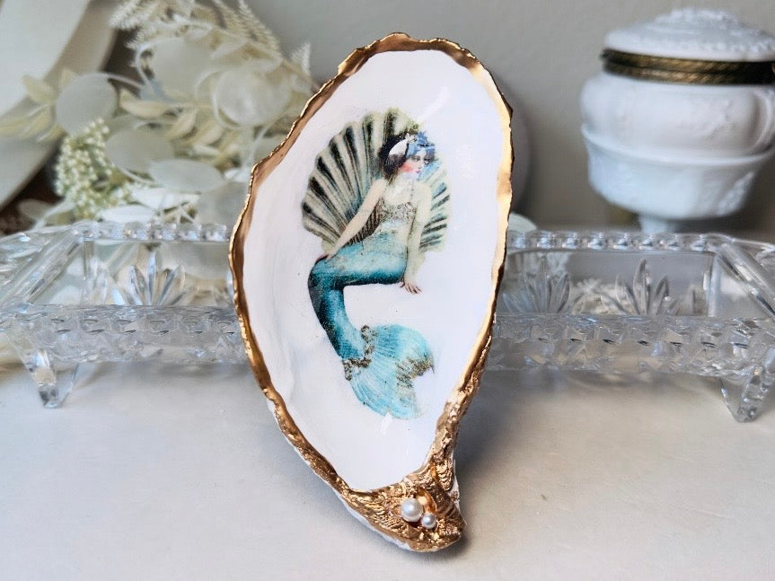 A decorative shell-shaped dish featuring a painted mermaid with a blue tail and gold-trimmed edges, displayed on a light surface with glass and white decorative objects in the background.