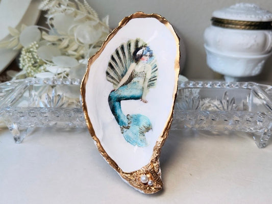 A decorative shell-shaped dish featuring a painted mermaid with a blue tail and gold-trimmed edges, displayed on a light surface with glass and white decorative objects in the background.
