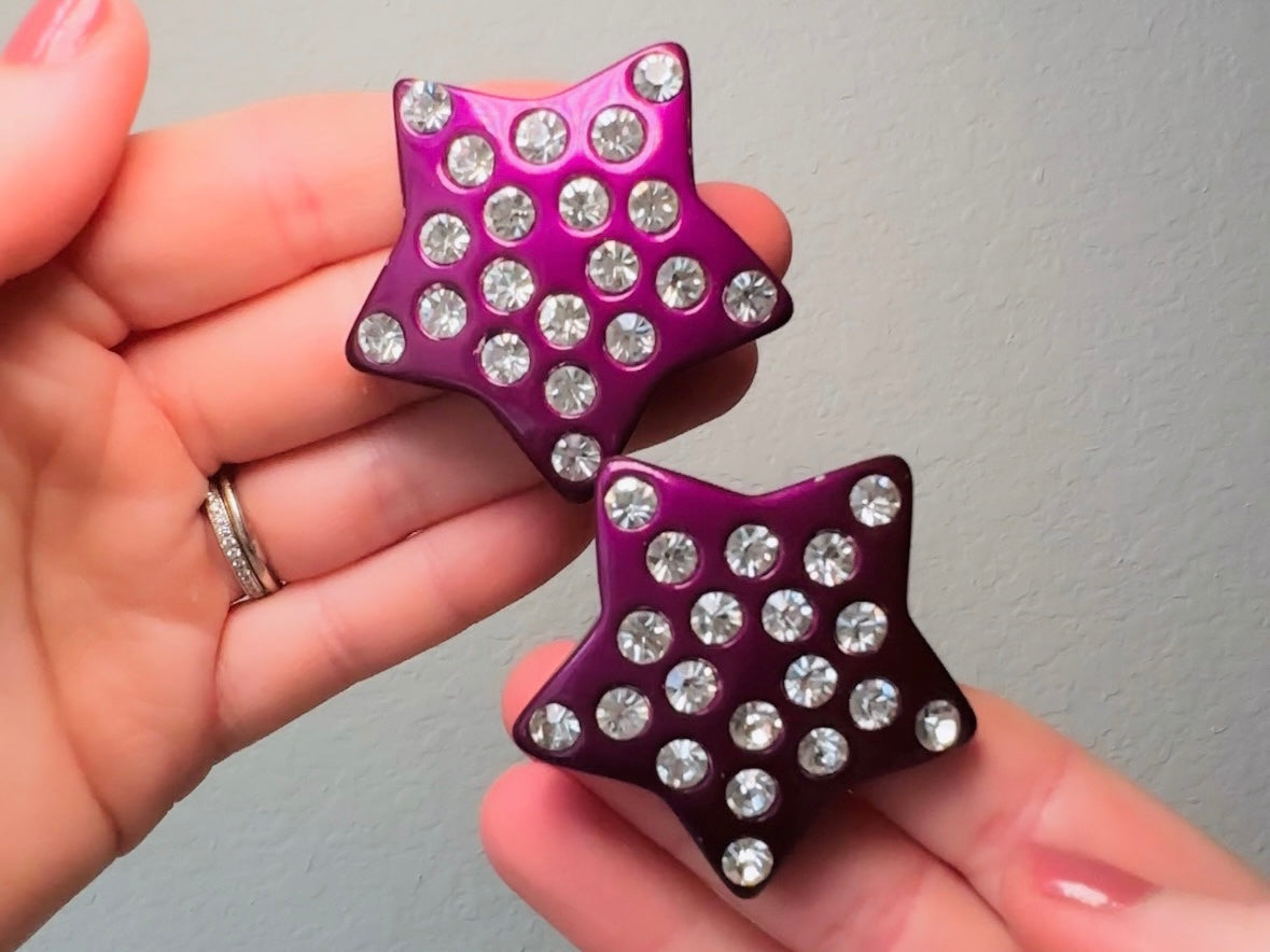 These oversized vintage purple star earrings are a highly collectible pair of 1980s statement earrings. These big and bold star earrings are a stunning shade of vibrant metallic magenta purple lucite with diamond rhinestone accents. Huge vintage star clip ons with major rockstar vibes. A fun 80s pair of unique earrings that you never knew you needed so badly.
