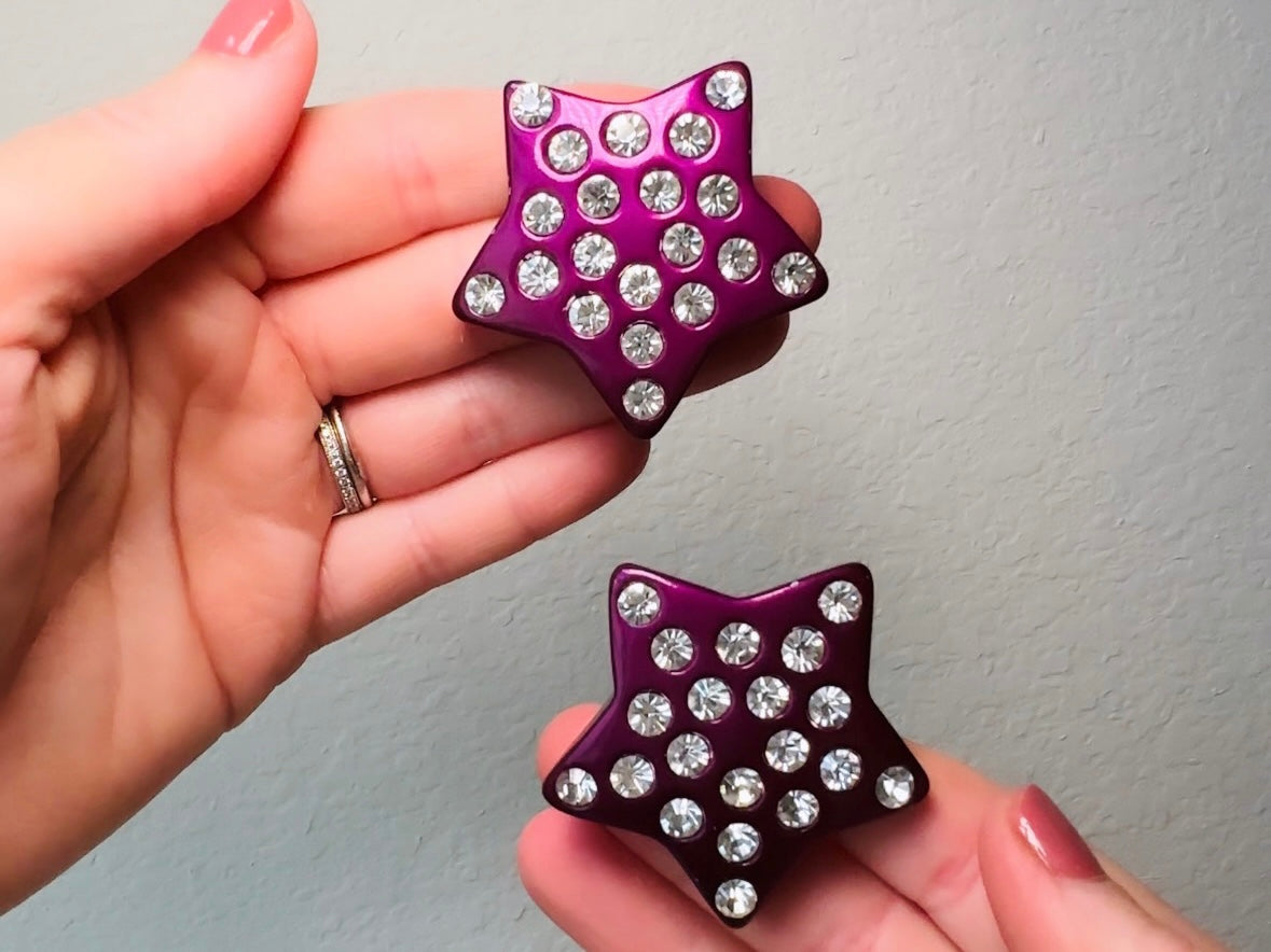 These oversized vintage purple star earrings are a highly collectible pair of 1980s statement earrings. These big and bold star earrings are a stunning shade of vibrant metallic magenta purple lucite with diamond rhinestone accents. Huge vintage star clip ons with major rockstar vibes. A fun 80s pair of unique earrings that you never knew you needed so badly.