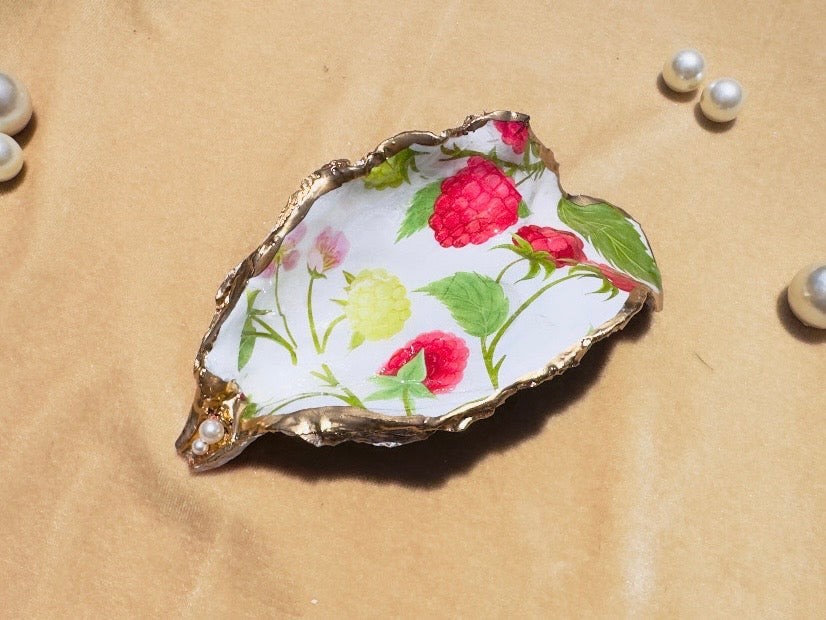 A decorative shell-shaped jewelry tray with a gold leaf-look rim and a white interior decorated with colorful floral and raspberry illustrations, placed on a tan textured surface with scattered pearl-like beads.