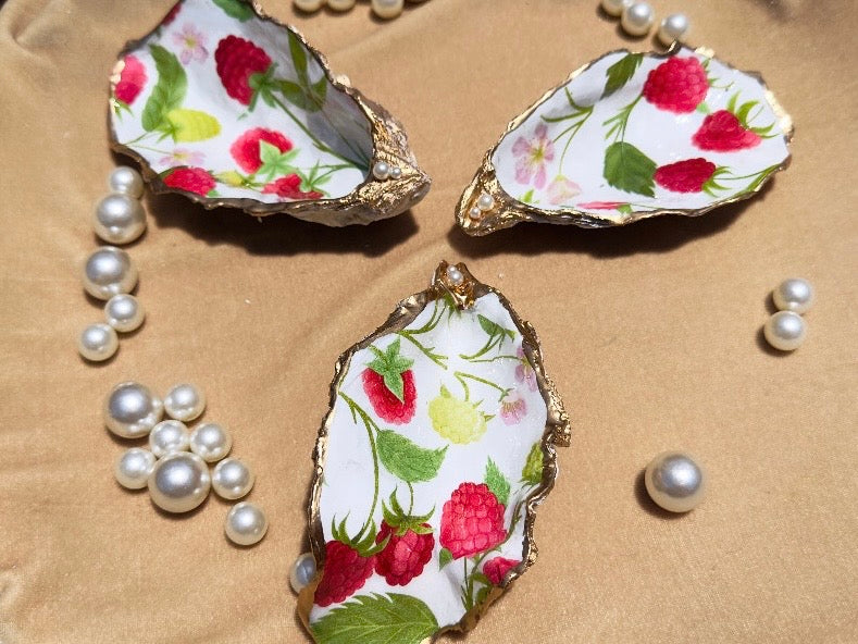 Three decorative oyster shell trays with gold-edged rims and a white background printed with red raspberries, green leaves, and small yellow flowers, arranged on a beige fabric surface with scattered faux pearls.