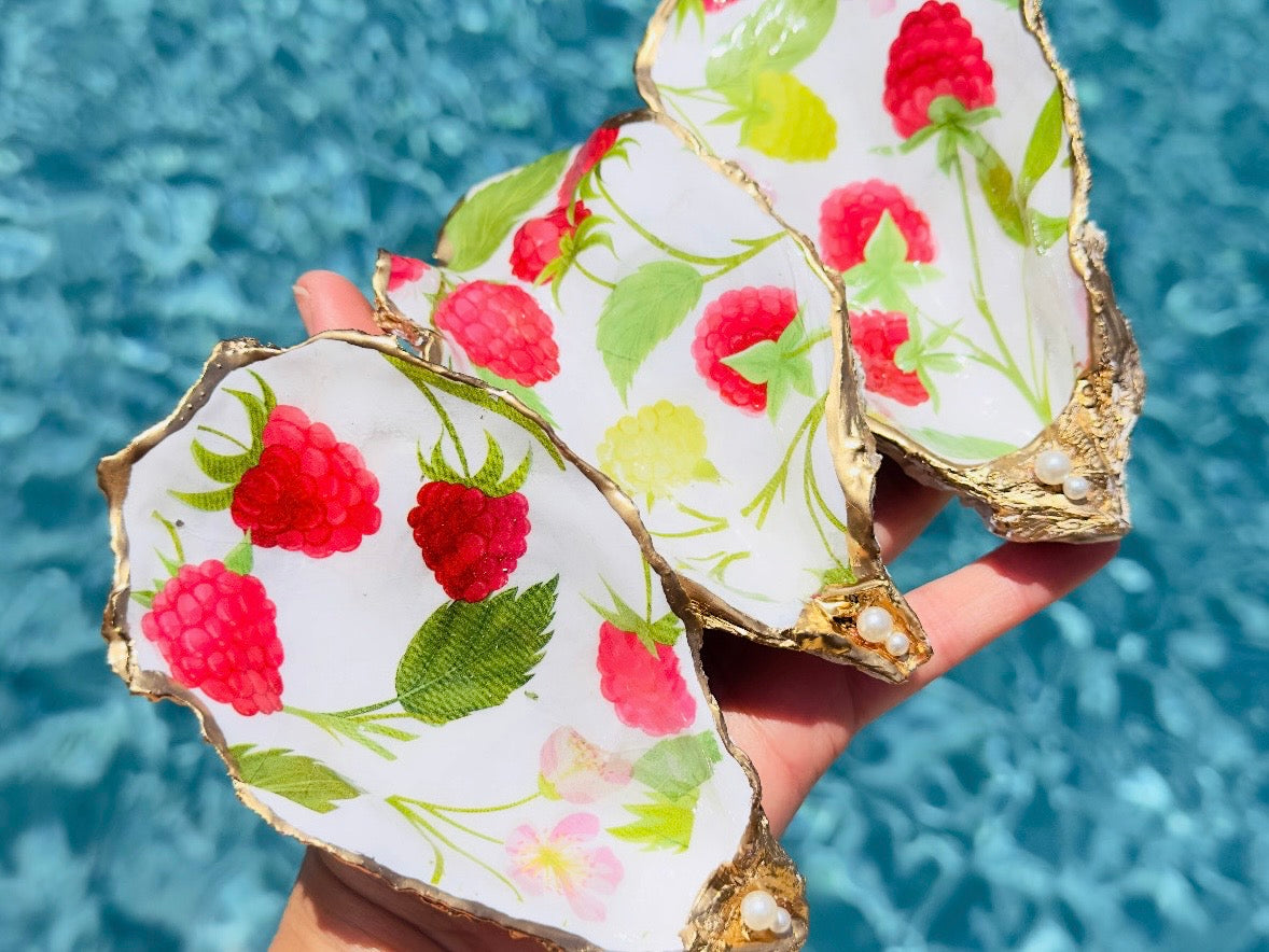 Three hand-painted oyster shells with a white background, decorated with red raspberries, green leaves, and gold-rimmed edges, held in a hand against a blue swimming pool background.