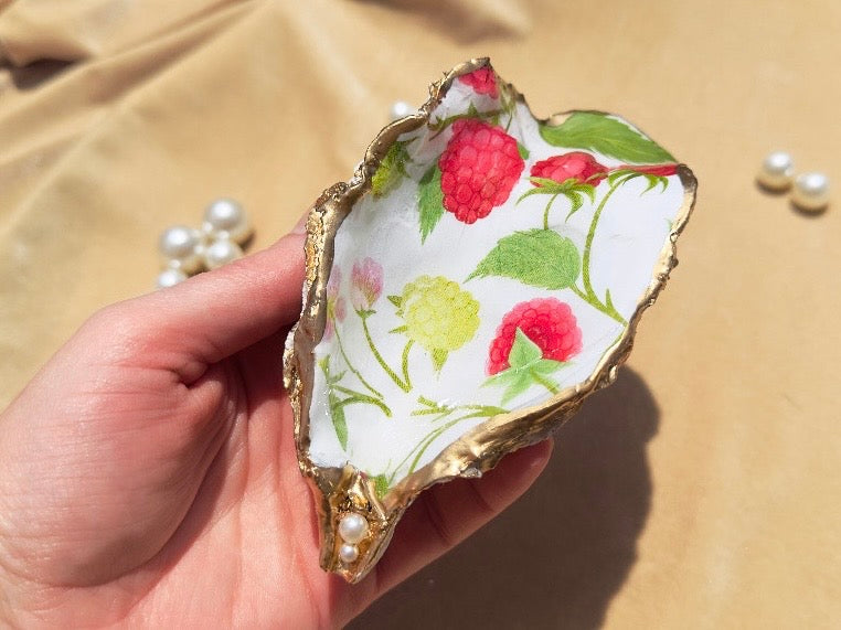 A hand holding a decorative oyster shell tray with a white interior printed with red raspberries, green leaves, and gold-toned edges, set against a beige fabric background with small pearl-like beads.