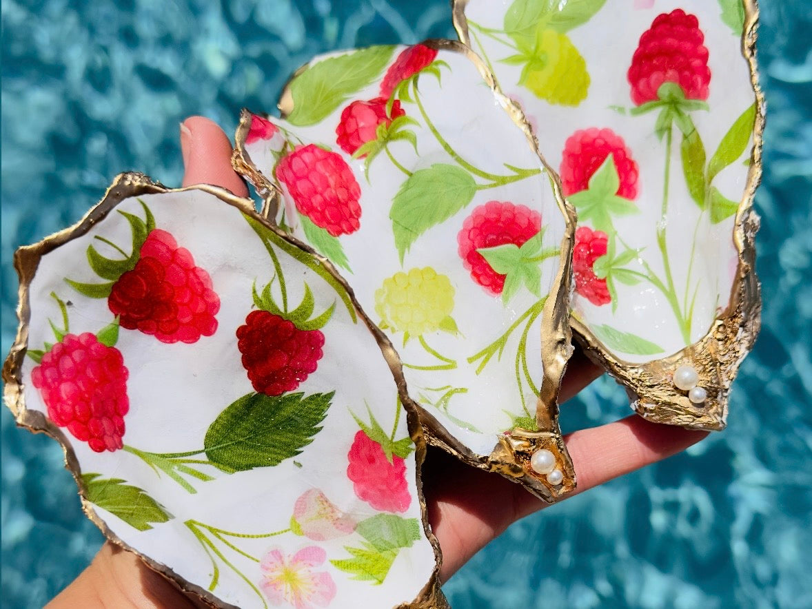 Three hand-painted oyster shells with raspberry and green leaf patterns, finished with gold-leaf edges and pearl accents, held in a hand against a blue water background.