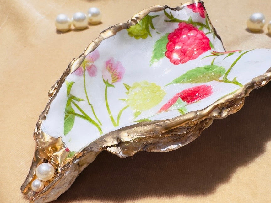A decorative shell-shaped dish with a white interior painted with pink and green floral motifs, bordered by metallic gold leaf, shown on a beige surface with small pearl-like beads nearby.