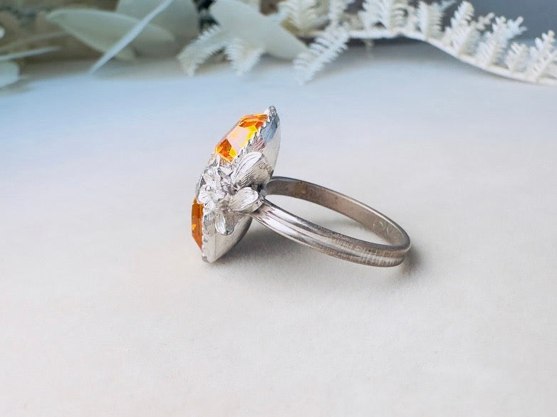 A silver ring featuring a marquise-cut orange gemstone and a floral cluster of clear stones on the band, displayed on a light surface with soft white decorative foliage in the background.