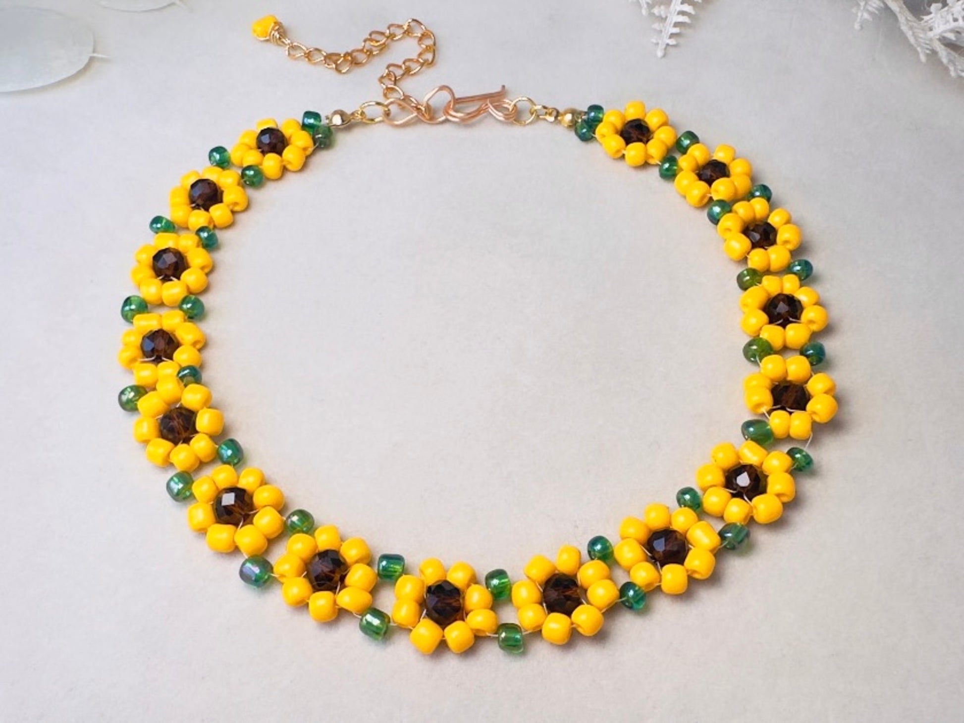 A handmade beaded necklace featuring yellow and green sunflower motifs with dark brown faceted centers, connected by a gold-tone chain extender and lobster clasp, displayed on a light gray surface.