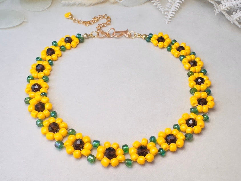 A beaded necklace featuring yellow flower petals with dark brown centers and green beads, arranged in a circle on a light gray background with subtle decorative white elements.