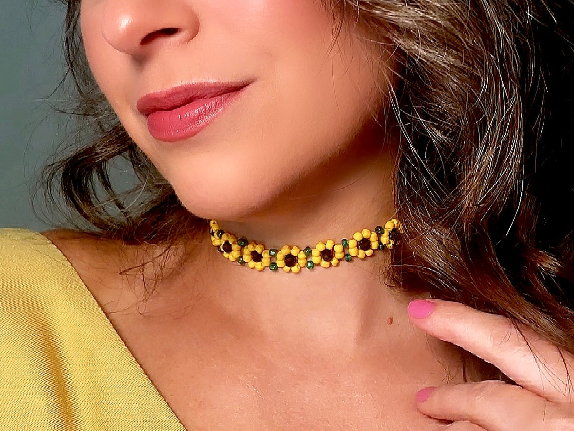 A close-up of a woman wearing a beaded choker necklace made of yellow beads with dark brown centers and small green accents, paired with a yellow top.