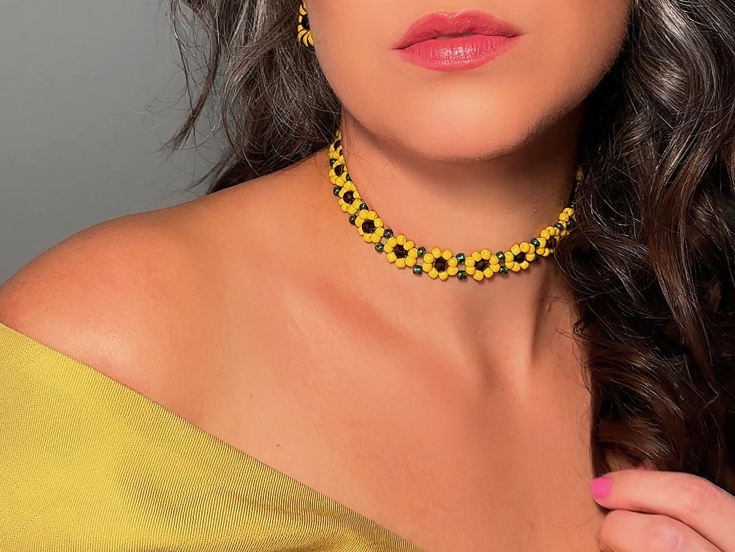 A close-up of a woman wearing a yellow and black beaded flower choker necklace, styled with a matching yellow top against a neutral gray background.