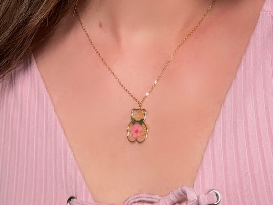 An adorable handmade teddy bear necklace modelled by a woman in a pink top. The necklace is made of 18kt gold and it has a cute teddy bear charm. The charm is clear with pink and yellow real pressed flowers. The bear is trimmed in gold and has a little gold bowtie.
