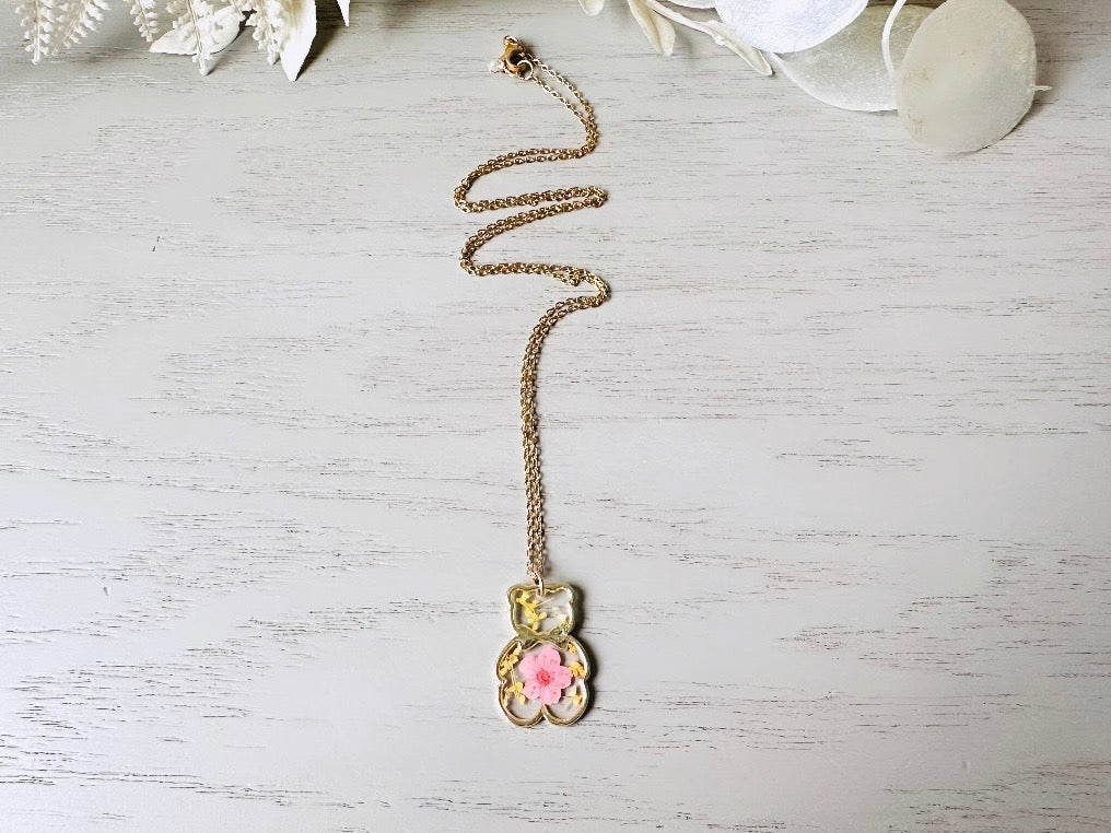 An adorable handmade teddy bear charm necklace. The necklace is made of 18kt gold and it has a cute teddy bear charm. The charm is clear with pink and yellow real pressed flowers. The bear is trimmed in gold and has a little gold bowtie.