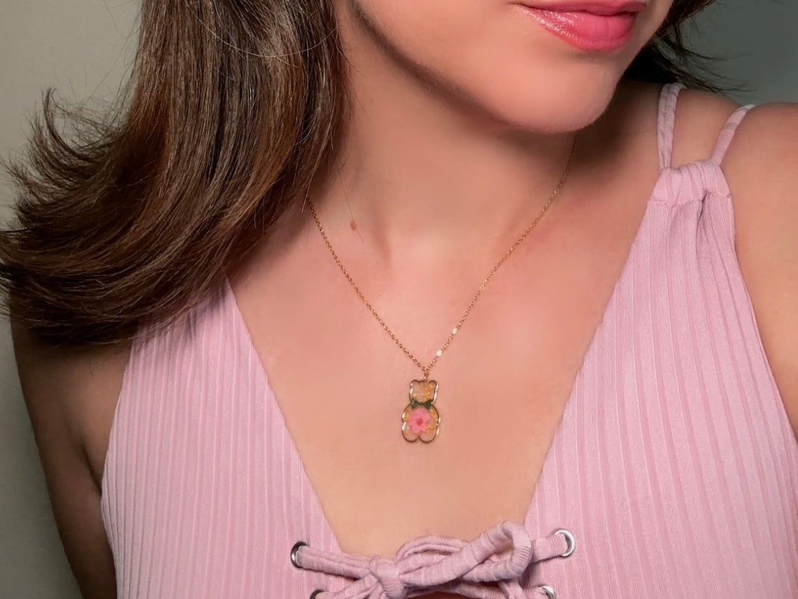 An adorable handmade teddy bear necklace modelled by a woman in a pink top. The necklace is made of 18kt gold and it has a cute teddy bear charm. The charm is clear with pink and yellow real pressed flowers. The bear is trimmed in gold and has a little gold bowtie.