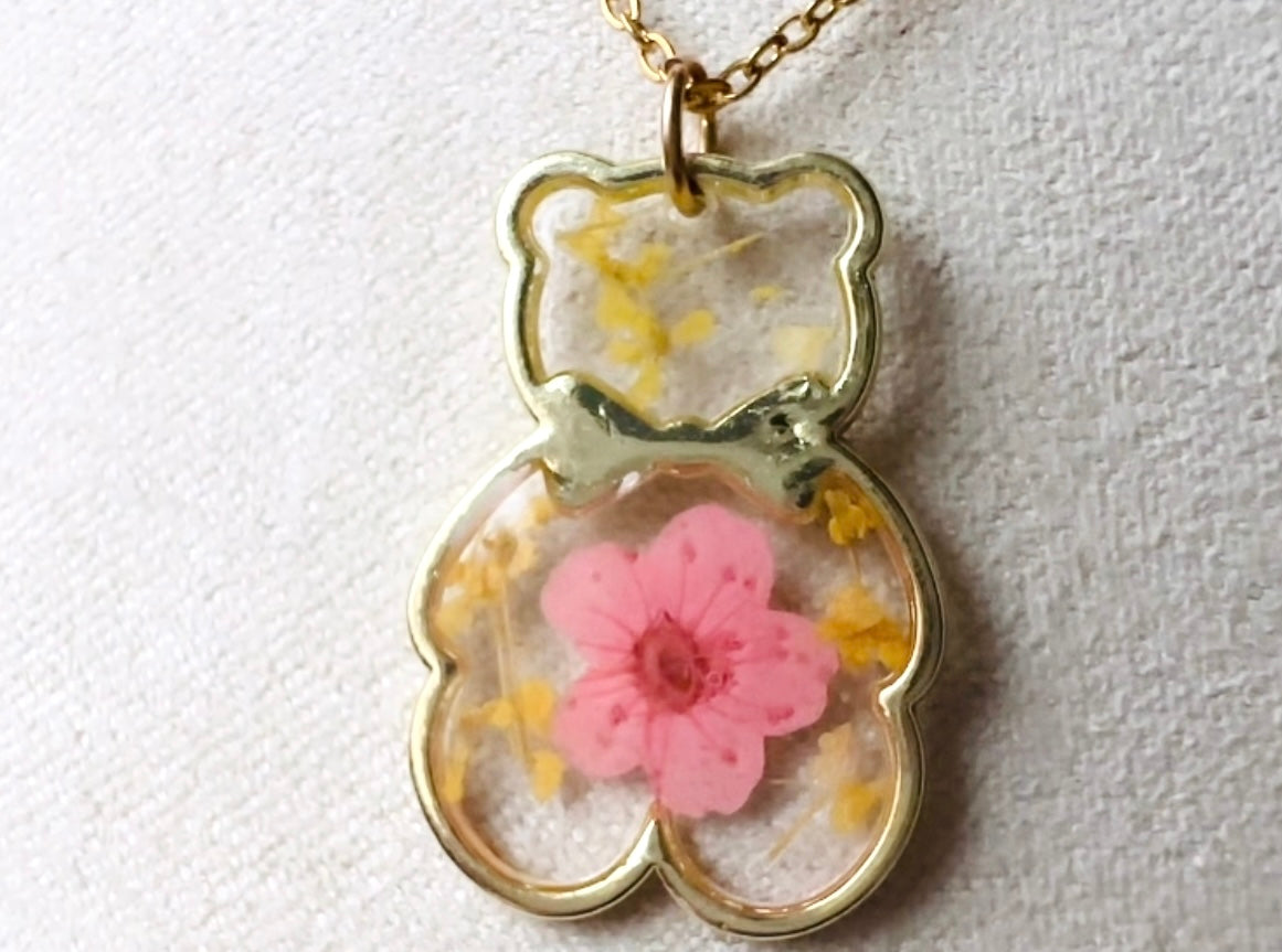 An adorable handmade teddy bear necklace modelled by a woman in a pink top. The necklace is made of 18kt gold and it has a cute teddy bear charm. The charm is clear with pink and yellow real pressed flowers. The bear is trimmed in gold and has a little gold bowtie.