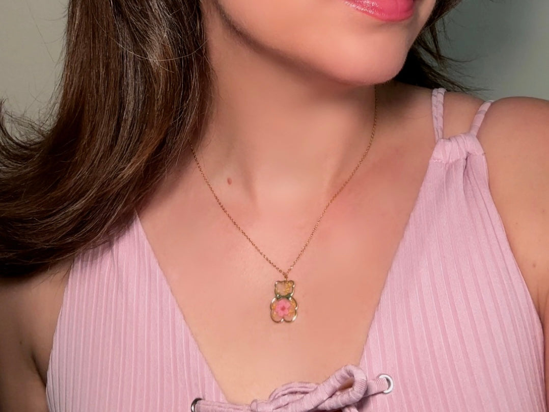 An adorable handmade teddy bear necklace modelled by a woman in a pink top. The necklace is made of 18kt gold and it has a cute teddy bear charm. The charm is clear with pink and yellow real pressed flowers. The bear is trimmed in gold and has a little gold bowtie.