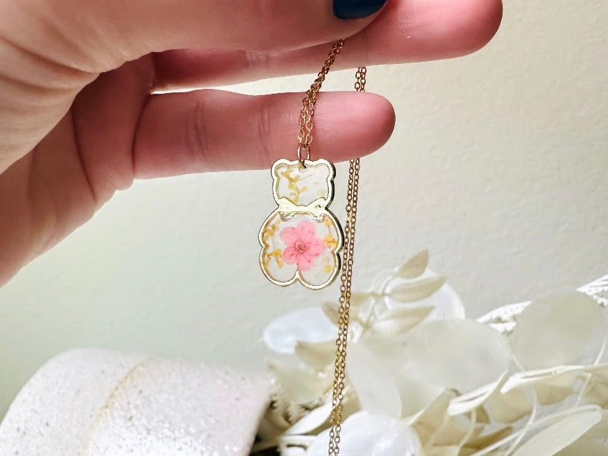 An adorable handmade teddy bear necklace. The necklace is made of 18kt gold and it has a cute teddy bear charm. The charm is clear with pink and yellow real pressed flowers. The bear is trimmed in gold and has a little gold bowtie.