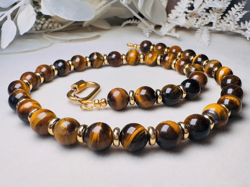 A beaded bracelet made of polished tiger's eye beads in brown and black with small gold spacer beads and a gold clasp, displayed on a light surface with white dried flowers in the background.