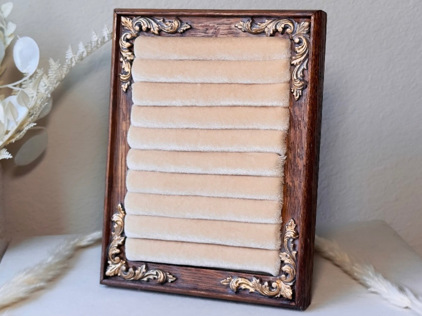 A rectangular jewelry organizer with a dark brown carved wooden frame and ornate gold corner details, holding ten horizontal beige velvet ring slots, shown standing on a light surface with dried floral decor in the background.