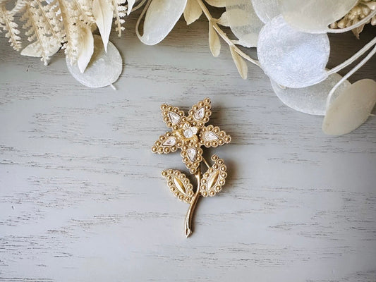 A beautiful vintage flower brooch by the brand 1928. The brooch is a single flower shape that is gold, with textured dotting all around each petal and leaf. There are diamond crystal rhinestones in a teardrop shape in the center of each flower petal and a round crystal in the center. The stem is a smooth shiny gold. The brooch is on the signature background of the shop Piggle and Pop, a light gray wood table with white floral background. 