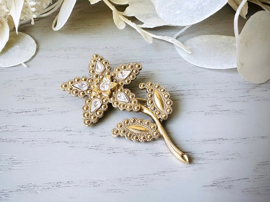 A beautiful vintage flower brooch by the brand 1928. The brooch is a single flower shape that is gold, with textured dotting all around each petal and leaf. There are diamond crystal rhinestones in a teardrop shape in the center of each flower petal and a round crystal in the center. The stem is a smooth shiny gold. The brooch is on the signature background of the shop Piggle and Pop, a light gray wood table with white floral background. 