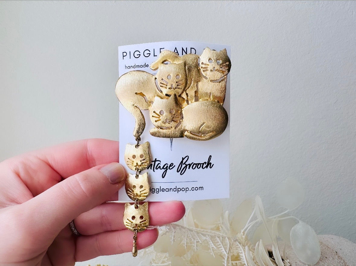This is a big gold vintage 1980s cat brooch featuring three cats huddled together and three cat heads dangling from their tail. The gold is a classic semi- matte shade with brushed detailing. It is a signed Ultra Craft piece in excellent vintage condition. 