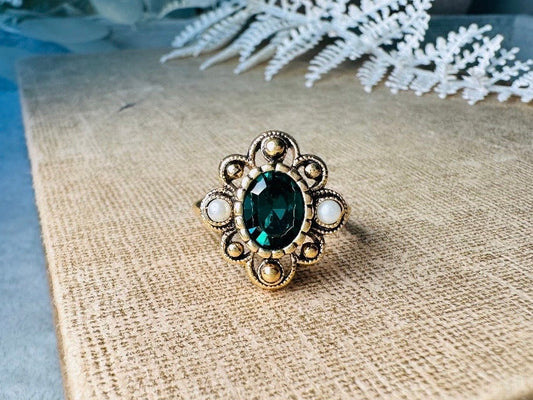 This vintage emerald green and pearl gold cocktail ring is such a pretty piece. Vintage Avon from the 1976 Versailles collection. Green, pearls and gold are a beautiful color combination. The center faceted oval green stone is surrounded in a raised filigree design that is full of vintage character, with two tiny pearl cabochon accents. Signed in excellent vintage condition.&nbsp;