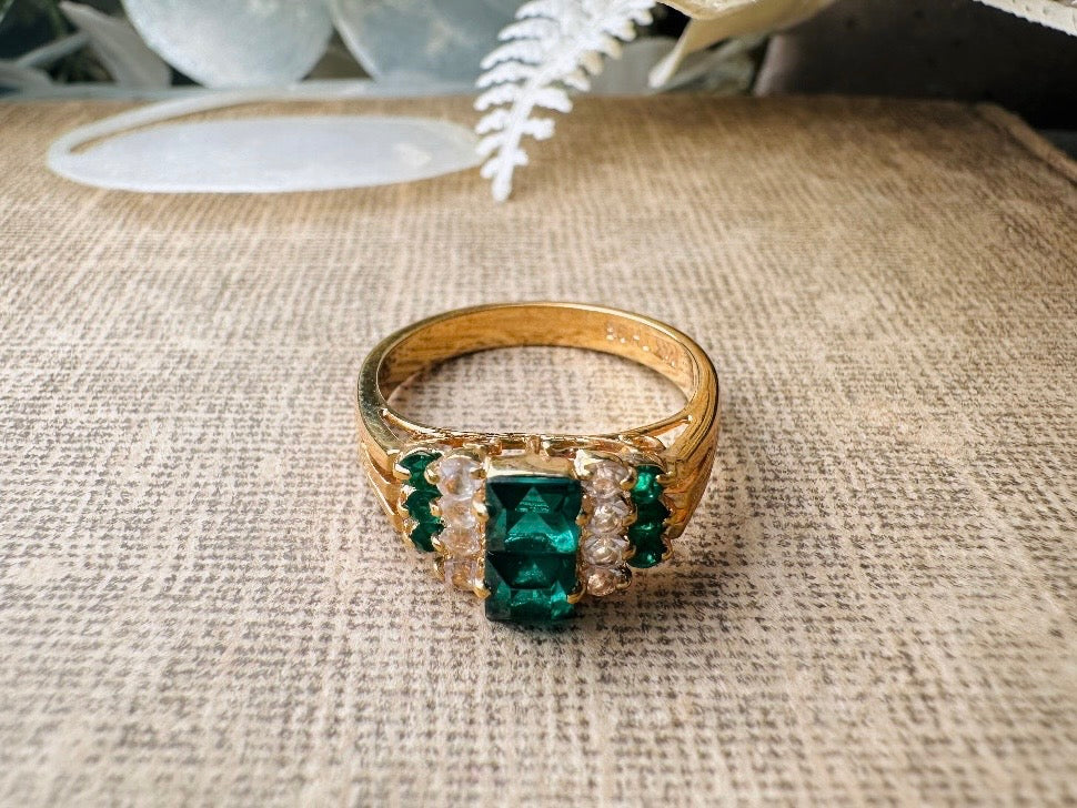 This vintage cocktail ring from Piggle and Pop features a stunning art deco design with brilliant emerald green stacked princess cut rhinestones flanked with dazzling cubic zirconias and tiny green stones. Prong set in a 14K gold-plated base, this fitted size 7 ring is a beautiful and timeless vintage piece. The glass crystals catch the light beautifully and are full of sparkle.