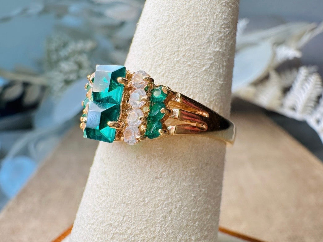 This vintage cocktail ring from Piggle and Pop features a stunning art deco design with brilliant emerald green stacked princess cut rhinestones flanked with dazzling cubic zirconias and tiny green stones. Prong set in a 14K gold-plated base, this fitted size 7 ring is a beautiful and timeless vintage piece. The glass crystals catch the light beautifully and are full of sparkle.