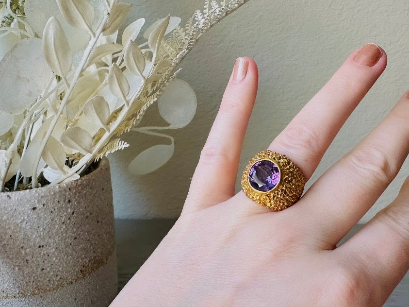 A vintage 1970s cocktail ring, that is gold and purple, The gold band has a raised textured gold nugget surface with a beautiful faceted round cut vibrant amethyst purple in the center. Worn on model for size reference.