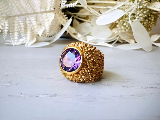 A vintage 1970s cocktail ring, that is gold and purple, The gold band has a raised textured gold nugget surface with a beautiful faceted round cut vibrant amethyst purple in the center.