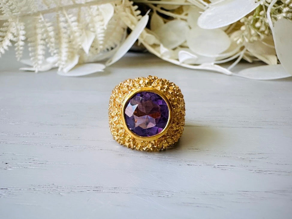 A vintage 1970s cocktail ring, that is gold and purple, The gold band has a raised textured gold nugget surface with a beautiful faceted round cut vibrant amethyst purple in the center.