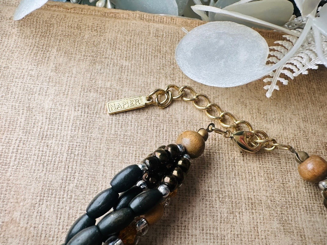 This vintage Napier necklace is a beautiful beaded twisted multistrand choker. Featuring six beaded strands in varying shapes and sizes composed of beads in wood, glass, acrylic, this bold necklace is a fabulous addition to your jewelry box. The beaded strands connect with a gold extension chain finished with a beautiful stamped Napier tag. 