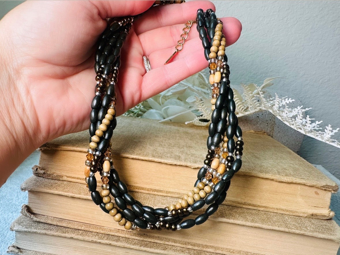 This vintage Napier necklace is a beautiful beaded twisted multistrand choker. Featuring six beaded strands in varying shapes and sizes composed of beads in wood, glass, acrylic, this bold necklace is a fabulous addition to your jewelry box. The beaded strands connect with a gold extension chain finished with a beautiful stamped Napier tag. Earthy black, brown and cream wood paired with gold, bronze and clear crystals- this necklace pairs beautifully with a variety of earrings and bracelets. Minimal wear in