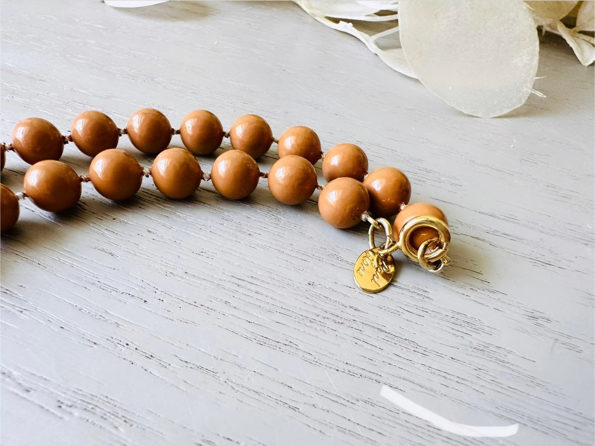 A vintage Park Lane designer necklace. The necklace is composed of 6mm caramel round beads with 5 bigger rondelle beads in the center with tiny gold spacer beads between. The necklace is on a light gray wood table with a floral background showing the signed Park Lane hang tag and clasp.