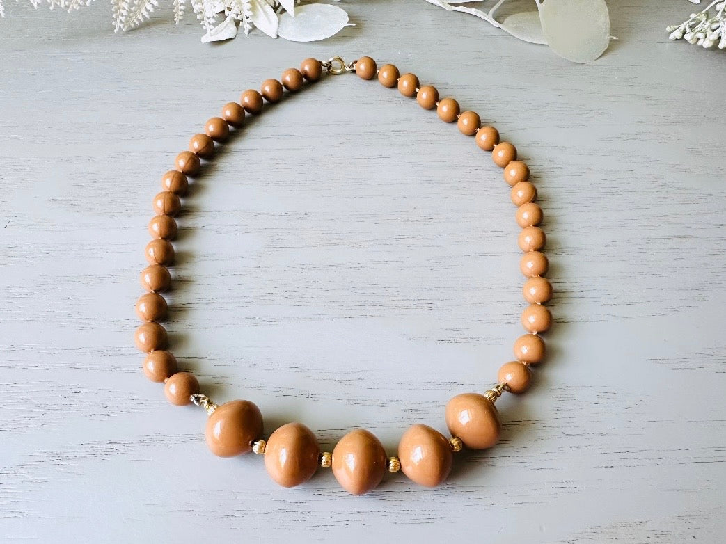 A vintage Park Lane designer necklace. The necklace is composed of 6mm caramel round beads with 5 bigger rondelle beads in the center with tiny gold spacer beads between. The necklace is on a light gray wood table with a floral background.